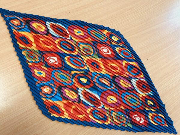 A vibrant kaleidoscope-patterned scarf with bold circular designs in red, blue, and orange, featuring a pleated texture, displayed on a wooden surface.
