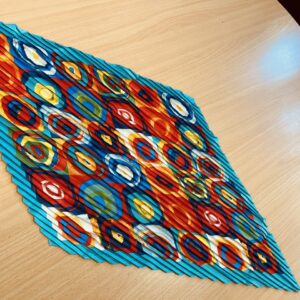 A bold kaleidoscope-patterned scarf with circular designs in red, blue, yellow, and orange, featuring a pleated texture, displayed on a wooden surface.