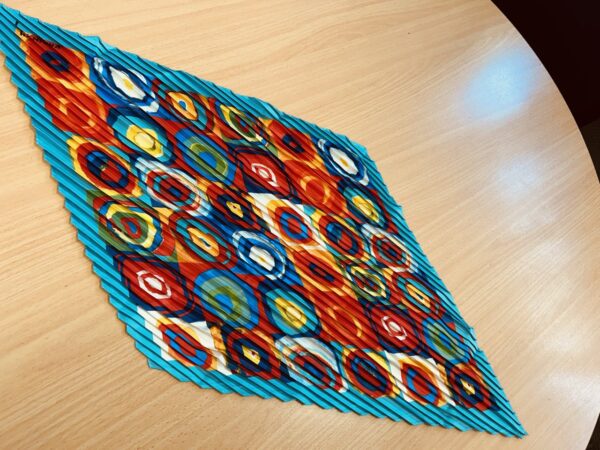 A bold kaleidoscope-patterned scarf with circular designs in red, blue, yellow, and orange, featuring a pleated texture, displayed on a wooden surface.