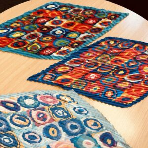 A bold kaleidoscope-patterned scarf with circular designs in red, blue, yellow, and orange, featuring a pleated texture, displayed on a wooden surface.