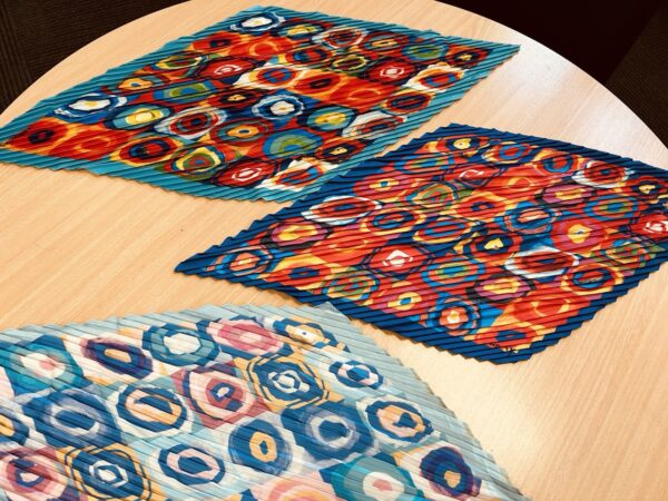 A bold kaleidoscope-patterned scarf with circular designs in red, blue, yellow, and orange, featuring a pleated texture, displayed on a wooden surface.