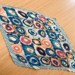A bold kaleidoscope-patterned scarf with circular designs in red, blue, yellow, and orange, featuring a pleated texture, displayed on a wooden surface.